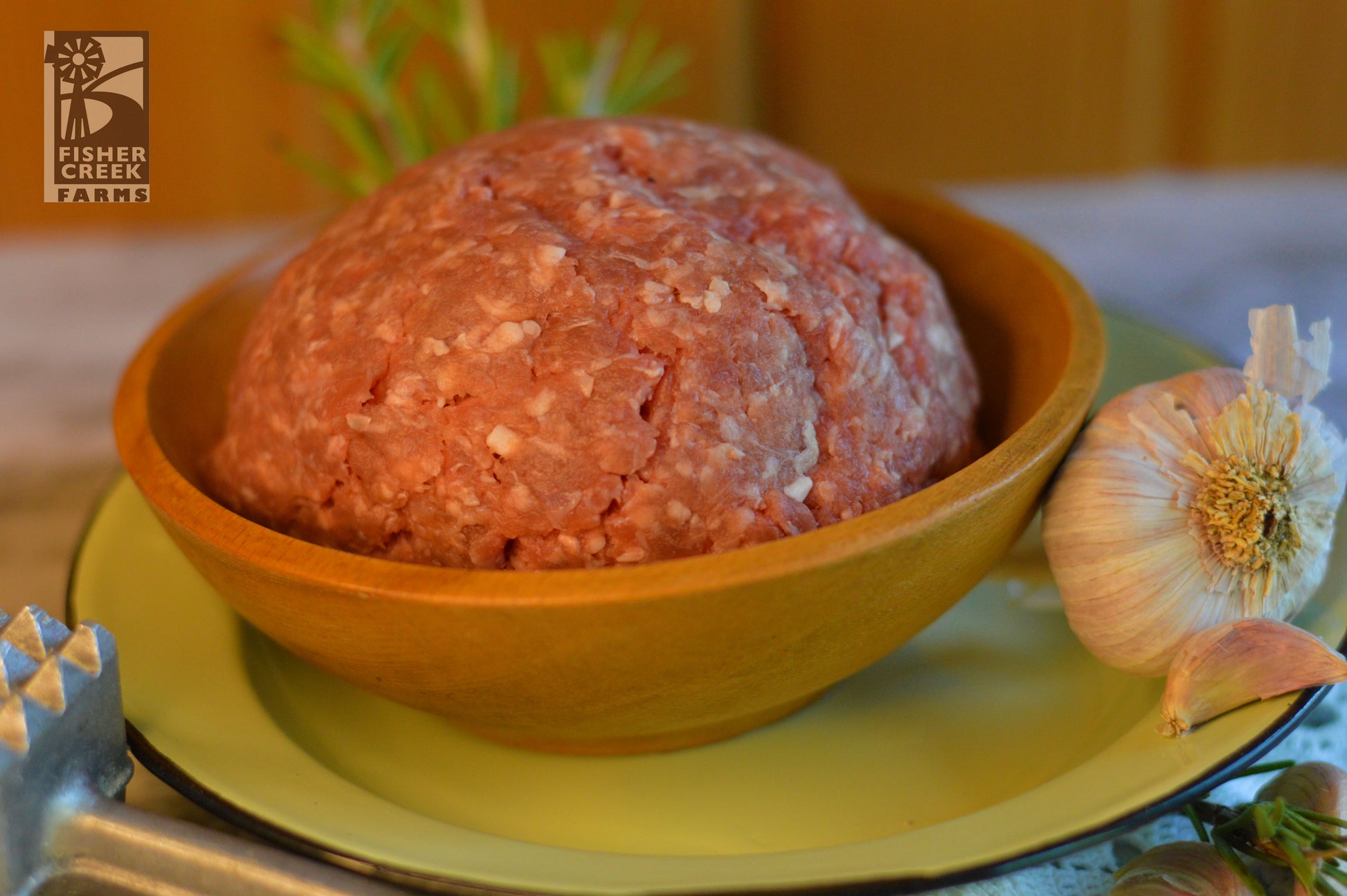 Bowl of ground goat meat on a yellow plate with a garlic bulb and rosemary sprig, branded 'Fisher Creek Farms'.