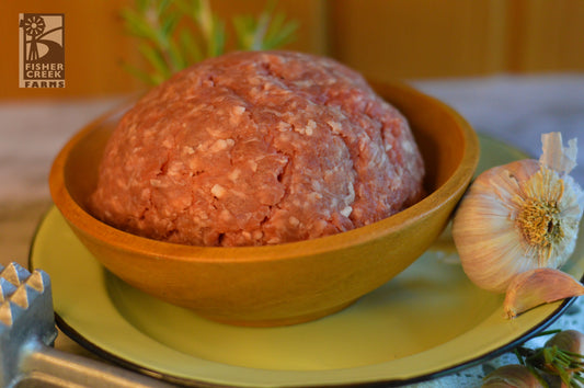 Bowl of ground goat meat on a yellow plate with a garlic bulb and rosemary sprig, branded 'Fisher Creek Farms'.