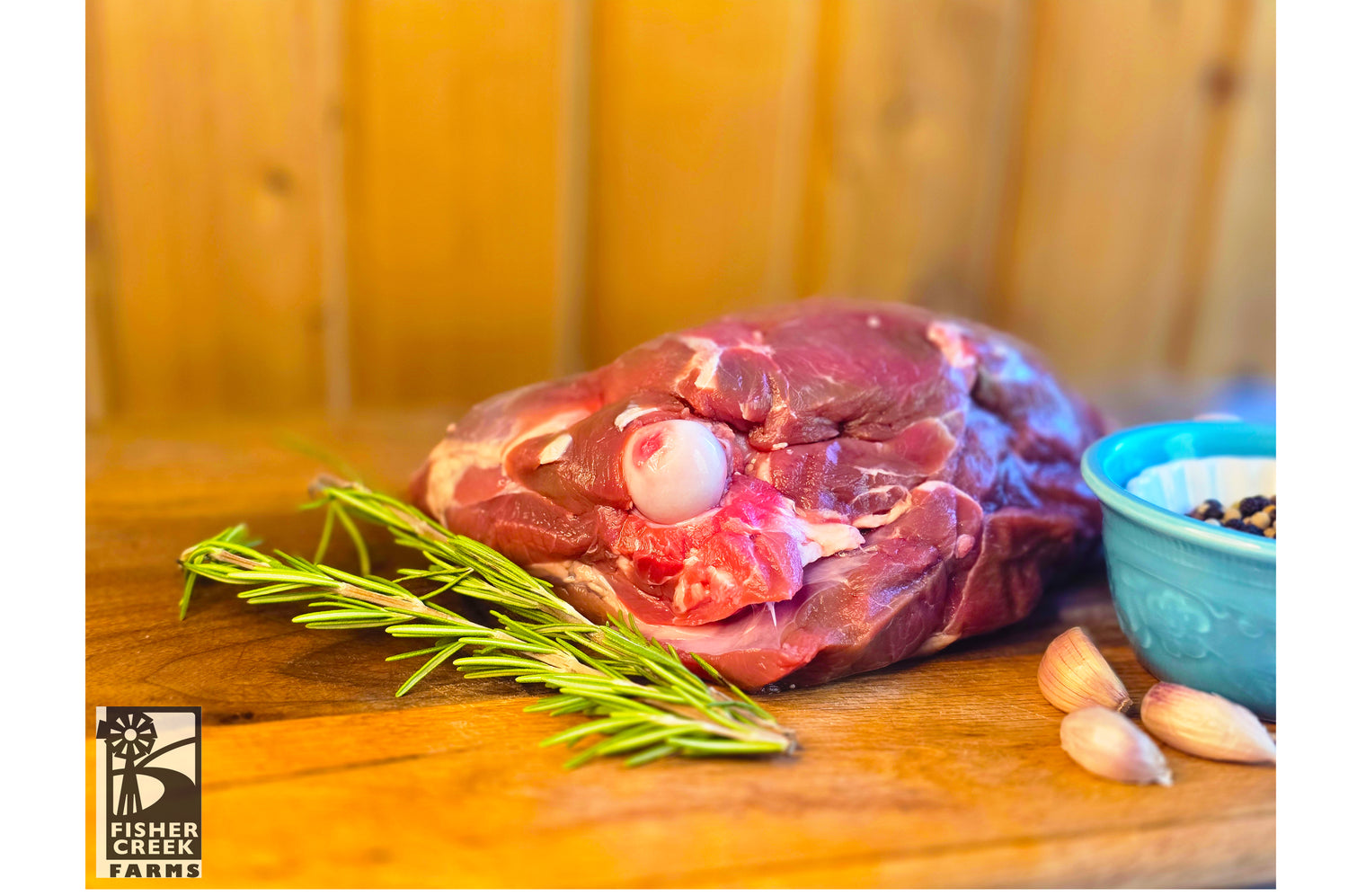 Raw leg of goat on a wooden cutting board with rosemary and garlic, branded 'Fisher Creek Farms'.