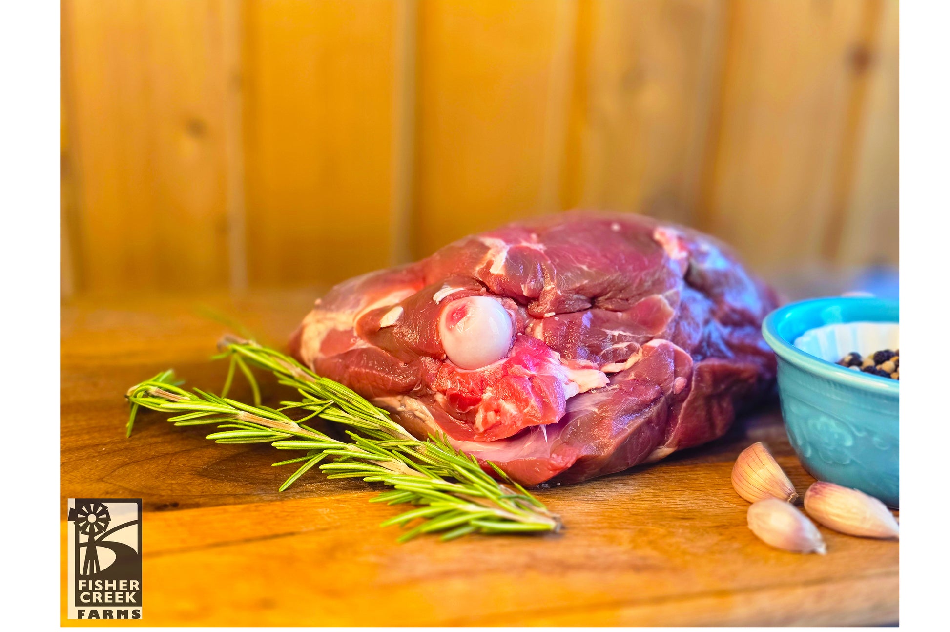Raw leg of goat on a wooden cutting board with rosemary and garlic, branded 'Fisher Creek Farms'.
