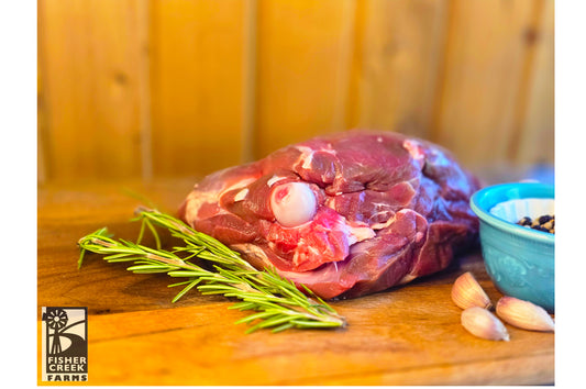 Raw leg of goat on a wooden cutting board with rosemary and garlic, branded 'Fisher Creek Farms'.